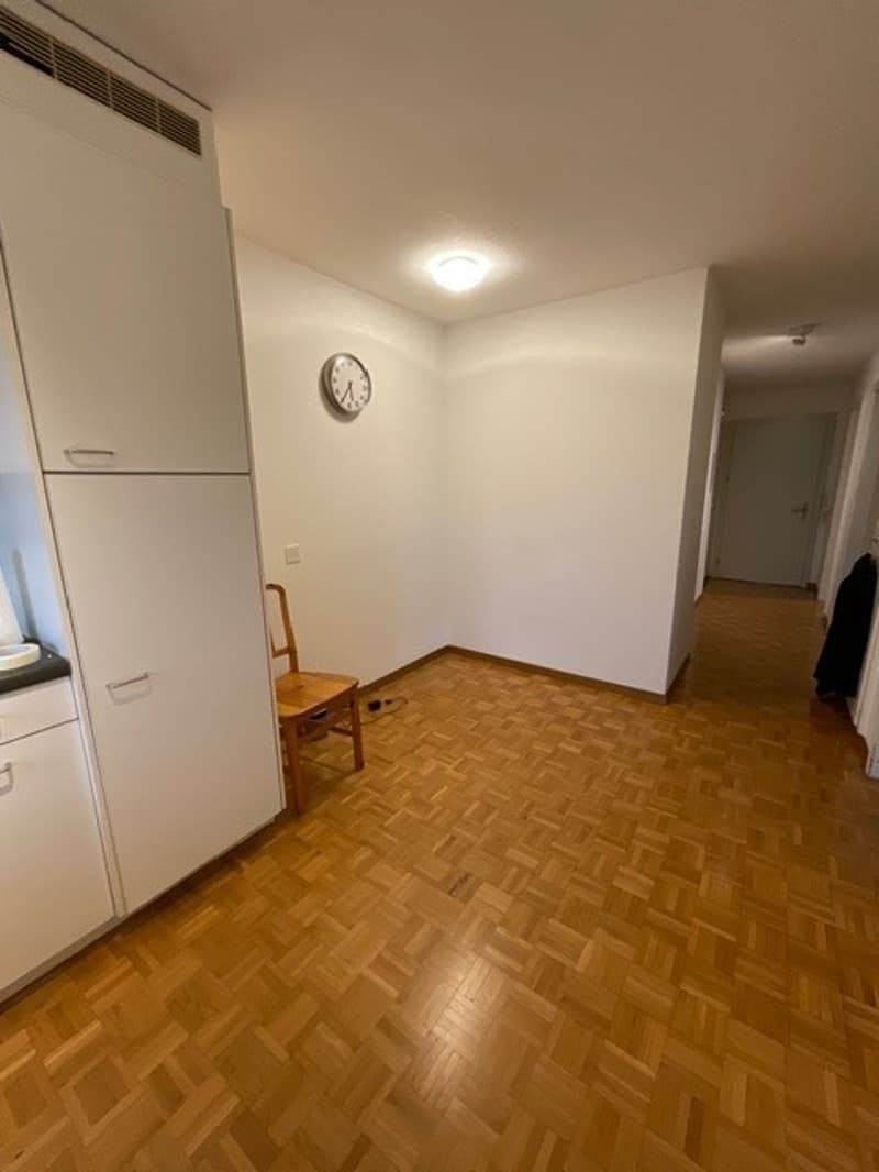 Empty room with wooden floor, white walls, wooden chair, white cabinets, clock on the wall, and a door leading to another room