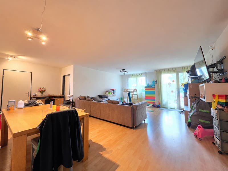 large living room with sofa, dining table, wooden floor, white walls, windows with curtains, TV, toys