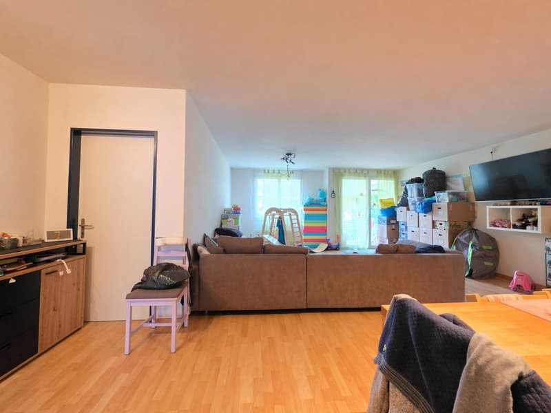 Living room with wooden floor, white walls, large brown sofa, wooden chair, wooden table, TV, shelves, and various items