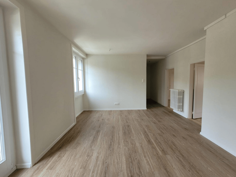 Spacious living room with light-colored hardwood flooring, white walls, and large windows providing natural light. The room appears to be empty, ready for furnishing and decorating.