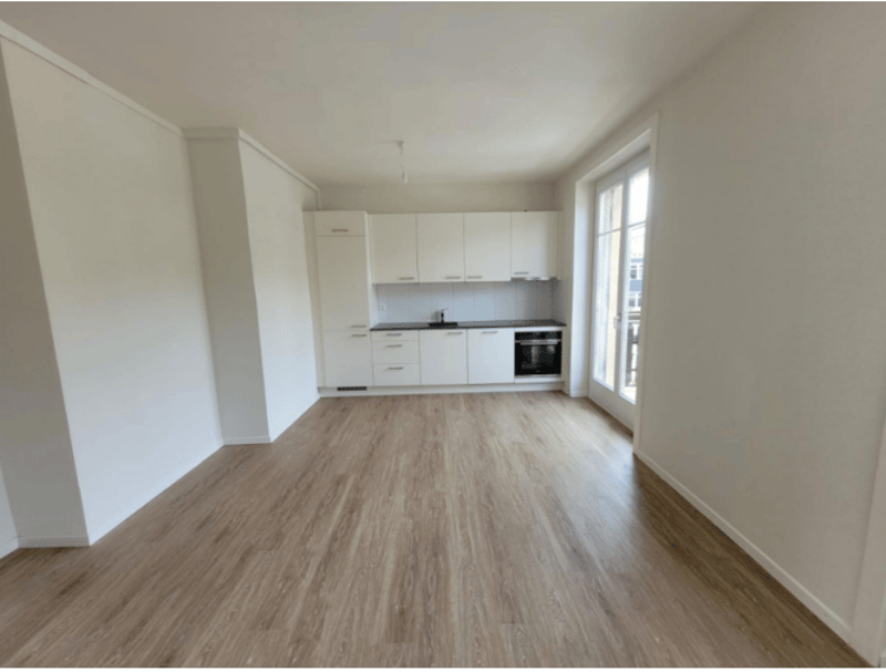An empty room with white walls, wooden floors, and a kitchen area with white cabinets and a black oven.