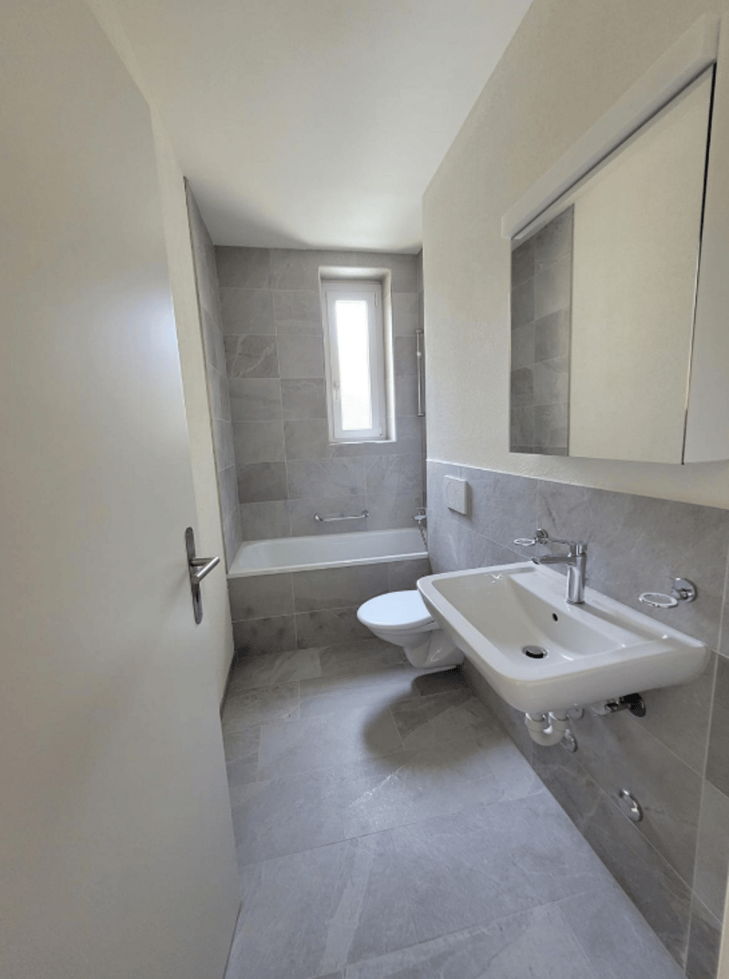 The image shows a modern bathroom with gray tile walls and floors, a white toilet, a white sink with a faucet, and a bathtub with a window.