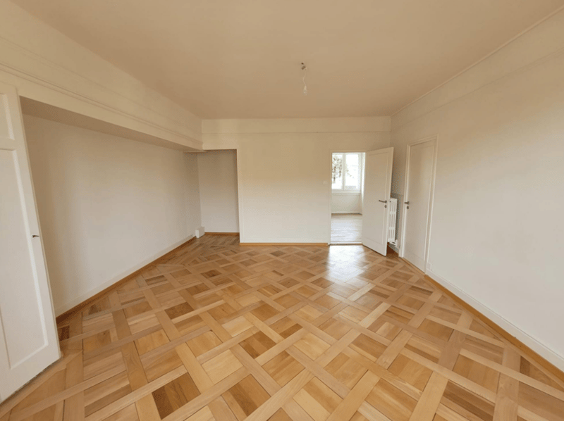 Spacious living room with hardwood parquet flooring, white walls, and a window providing natural light.