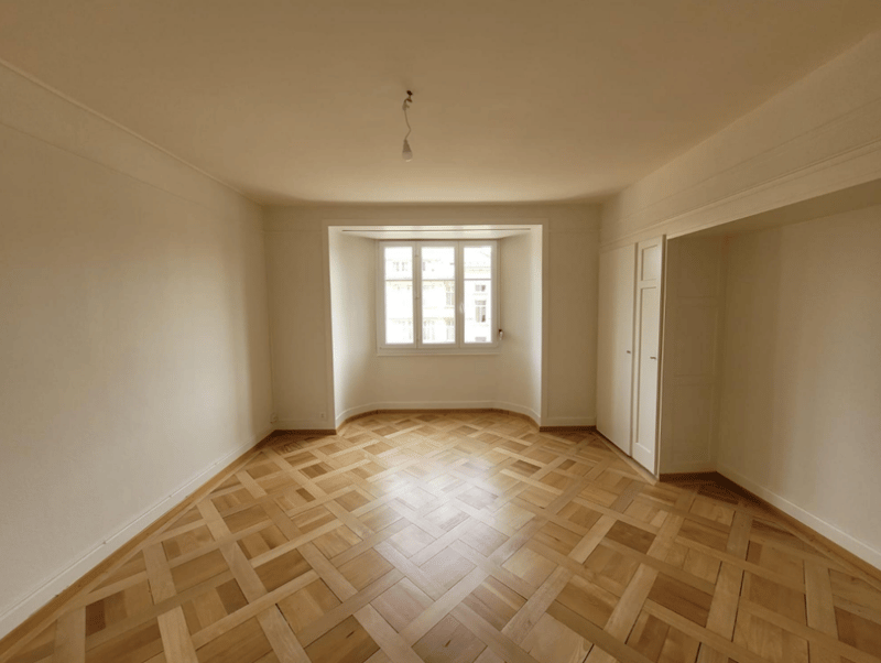 Spacious room with hardwood parquet flooring in a herringbone pattern, white walls, and a large window providing natural light.