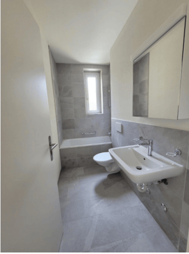 Bathroom with white bathtub, sink, toilet, and mirror. Gray tiled floor and walls.