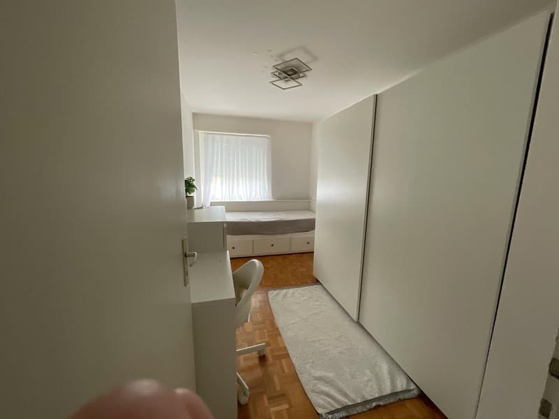 Empty room with white walls, wooden floor, desk, chair, rug, and a small bed with drawers