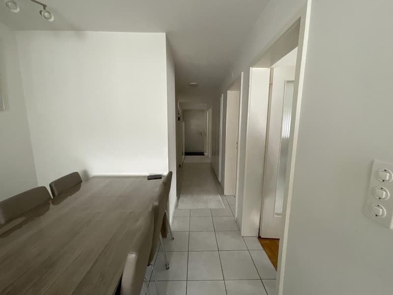 Dining room with a long table and chairs, hallway with white tiles, white walls, and doors.