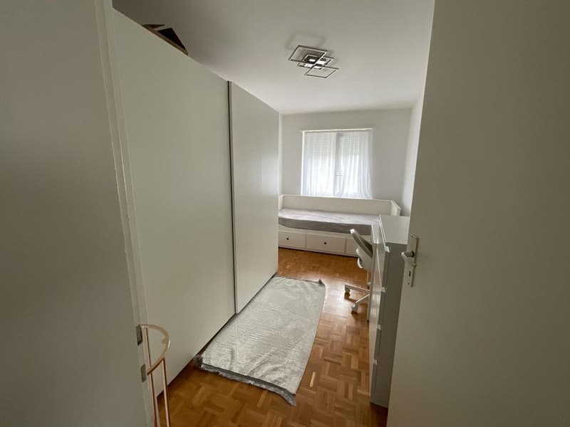 Empty room with wooden floor, white walls, a bed, desk, and a window with white curtains.