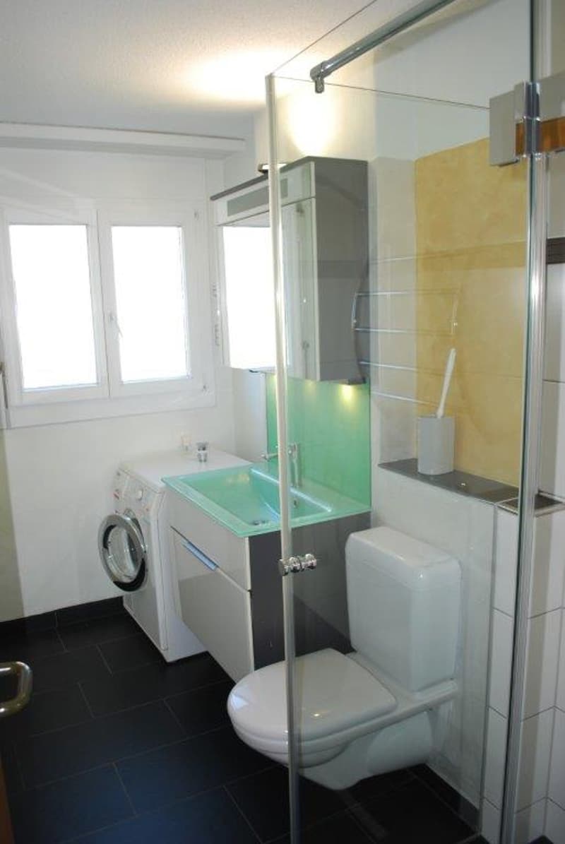 Modern bathroom with tiled floor, glass shower, white toilet, washing machine, sink, and large window