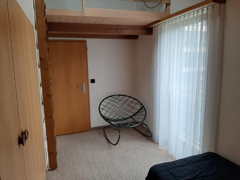 small room with wooden door, bed, chair, carpeted floor, white curtains