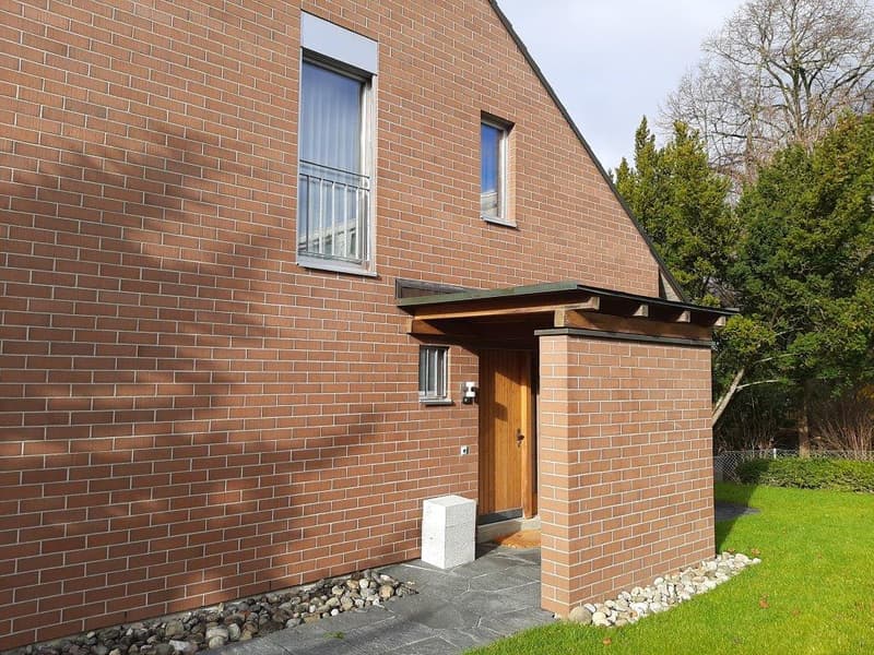 Red brick house, wooden door, two windows, small porch, gravel path, green lawn, trees