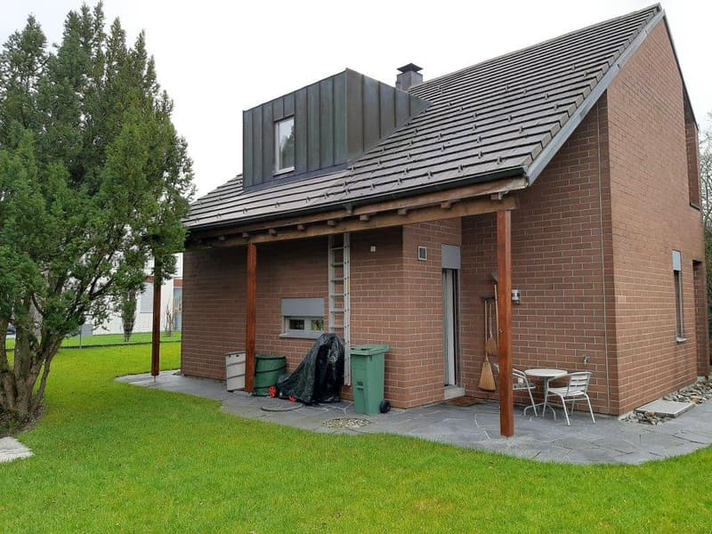 Single-story brick house, gray roof, wooden porch, garden, outdoor table and chairs, trash bins, ladder