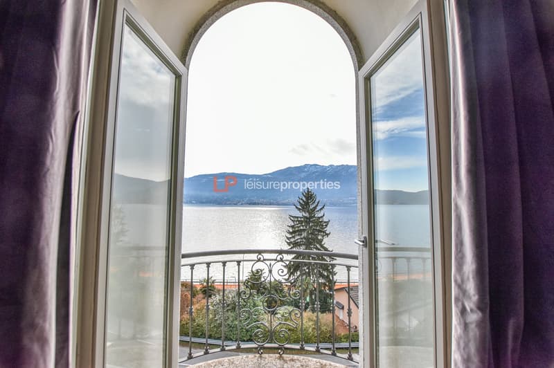 A room with an open balcony facing the lake with a picturesque view of the mountains, double glazed windows, and a decorative railing.