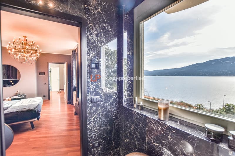 A room with a double bed, chandelier, wooden floor, large window with lake view, bedside table, mirror, and a window sill