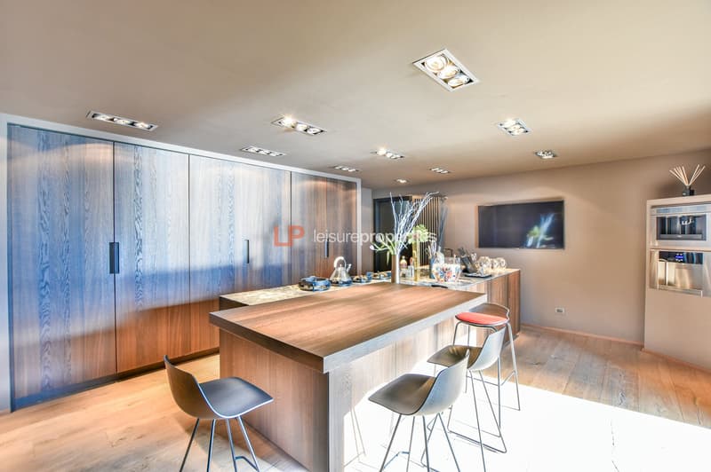 modern kitchen with central island, wooden cabinets, large countertop, four stools, gas stove, kettle, monitor on wall, integrated appliances