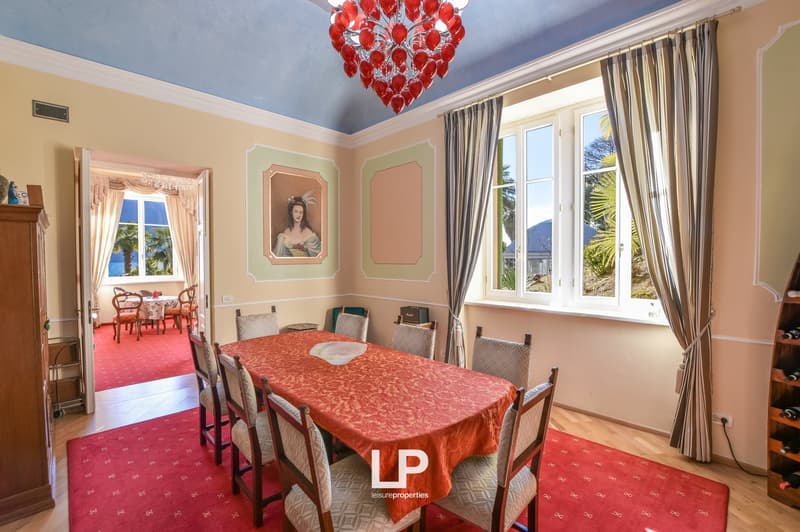 long dining table with 6 chairs, painting, chandelier, carpet, large windows, another room visible in the background