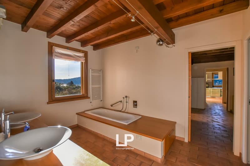 Spacious bathroom with wooden beamed ceiling, large window with scenic view, bathtub, and tiled floor.