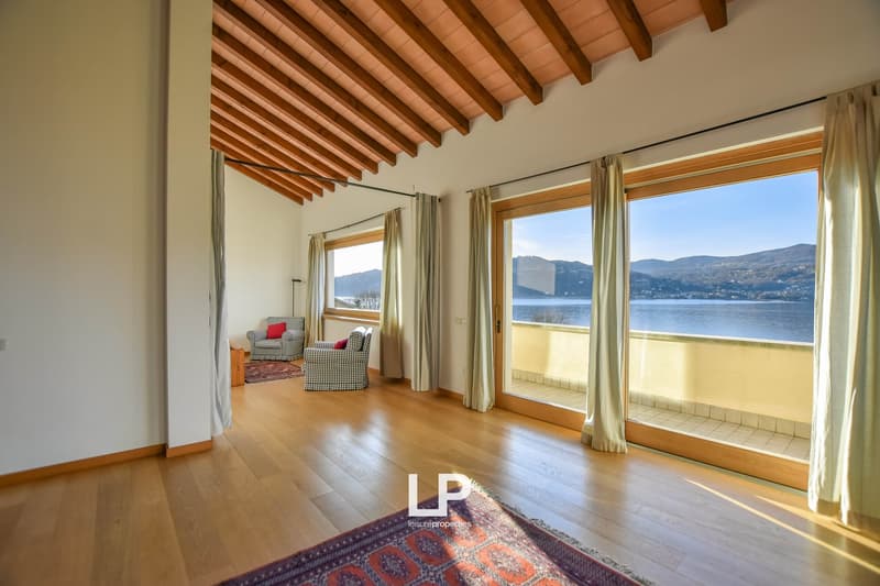 Spacious living room with wooden beamed ceiling, large windows offering a scenic view of the lake and mountains, hardwood flooring, and a cozy seating area with patterned armchairs.