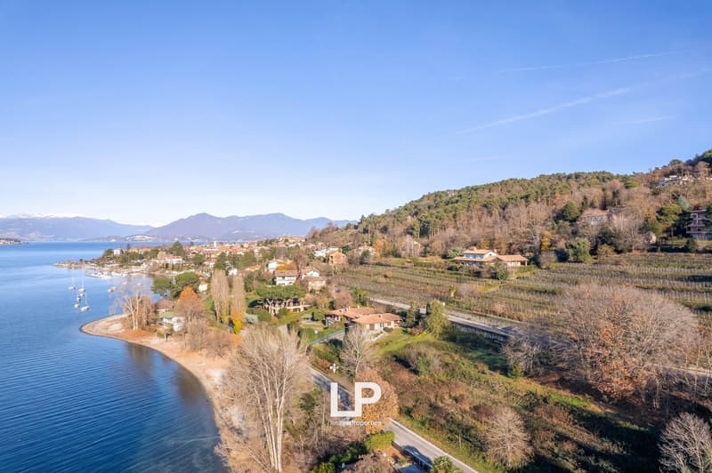 The image shows a scenic view of a lakeside town surrounded by mountains and hills. The town is situated along the shore of a large body of water, likely a lake. The landscape features a mix of residential buildings, vineyards, and forested areas. The sky