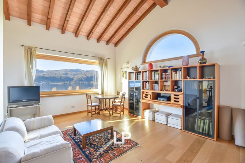 Spacious living room with wooden beamed ceiling, large windows overlooking a scenic lake view, wooden bookshelf, dining table, and sofa