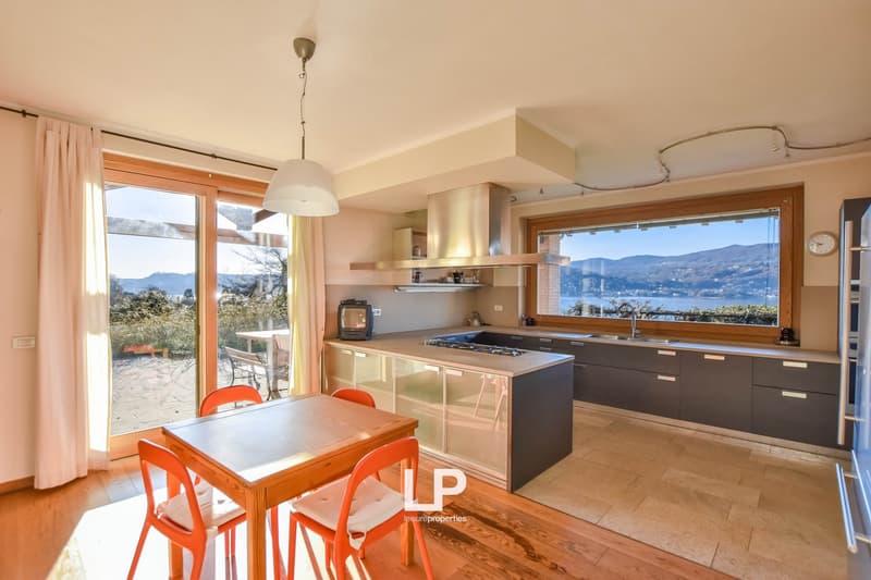 Open-concept kitchen and dining area with large windows offering a scenic view of the surrounding mountains and lake. The kitchen features modern dark cabinets, a gas stove, and a breakfast bar. The dining area has a wooden table and orange chairs. The ro