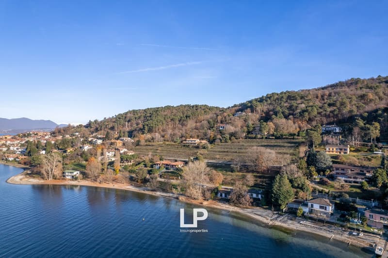 The image shows a scenic landscape with a lake or river in the foreground, surrounded by rolling hills and forests. There are several houses and buildings visible on the hillsides. The sky is clear and blue, creating a picturesque setting.