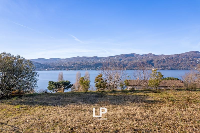 The image shows a scenic view of a lake surrounded by mountains and hills. There are trees and vegetation in the foreground, and a small building or structure can be seen in the distance. The sky is clear and blue, creating a peaceful and serene atmospher