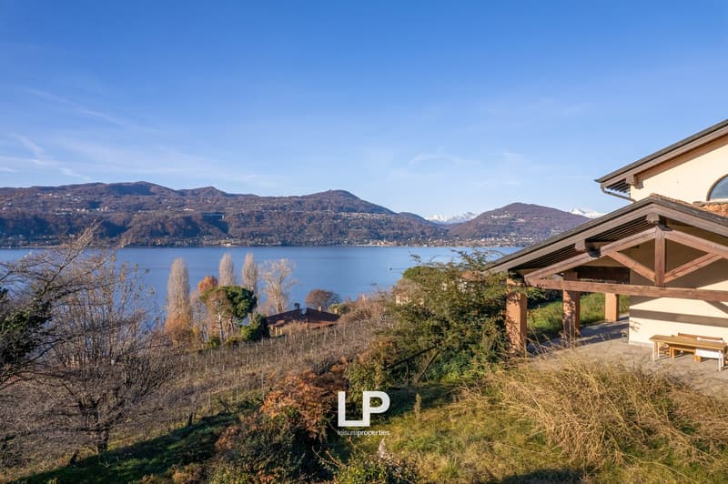 Two-story house with a wooden exterior, surrounded by lush greenery and overlooking a scenic lake and mountains in the distance. The house has a covered patio or balcony area with a view of the beautiful natural landscape.