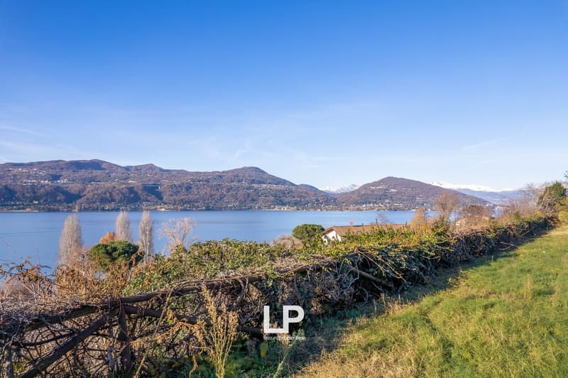 The image shows a scenic view of a lake surrounded by mountains and hills. The landscape includes a grassy area with trees and shrubs along the shoreline, with a few houses visible in the distance. The sky is clear and blue, creating a picturesque setting