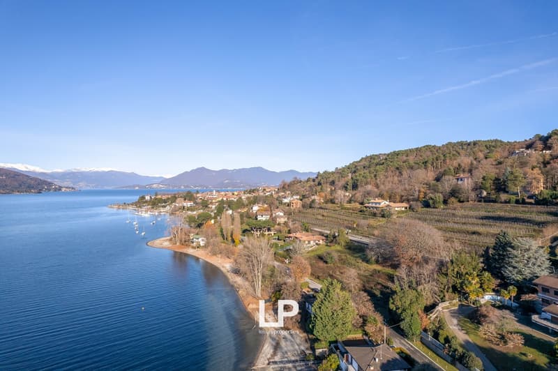 Aerial view of a lakeside town surrounded by mountains and hills, with vineyards and forests visible on the slopes. The town is situated along the shoreline of a large lake, with boats and docks visible in the water.