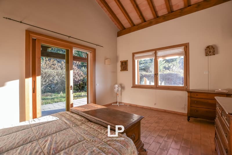 Spacious bedroom with wooden beamed ceiling, large windows overlooking a garden, wooden furniture including a dresser and a wooden table, and a sliding glass door leading to a balcony or terrace.