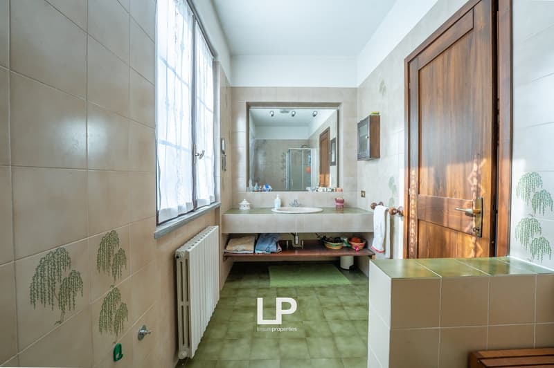 Bathroom with white tiles, green tiles on the floor, white sink, mirror, wooden door, radiator, towels, and a window with white curtains.