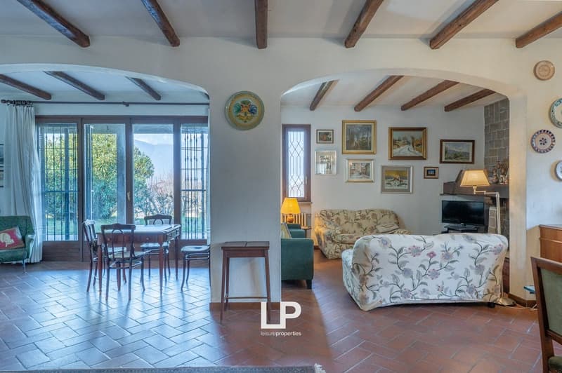 living room with dining area, wooden beams, brick floor, large windows, dining table, chairs, sofa, armchair, TV, fireplace, paintings on the wall