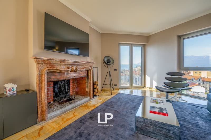 Spacious living room with a large ornate fireplace, hardwood floors, and large windows providing a view of the city. The room is furnished with a TV mounted above the fireplace, a tripod lamp, and a set of round stools or side tables.
