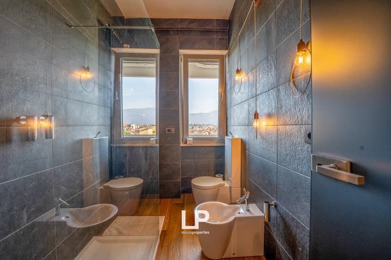 Spacious bathroom with large windows offering a scenic view, tiled walls in dark gray, modern fixtures including a toilet, bidet, and sink, and hanging light fixtures providing warm lighting.