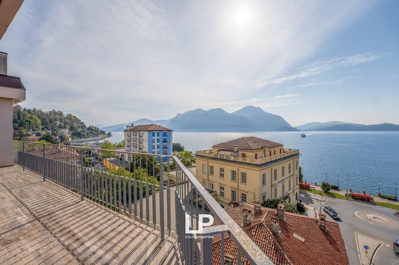 Balcony with a view of a lakeside town, surrounded by mountains. The image shows several multi-story buildings, including a historic-looking building with a red tile roof. There is also a boat on the lake visible in the distance.