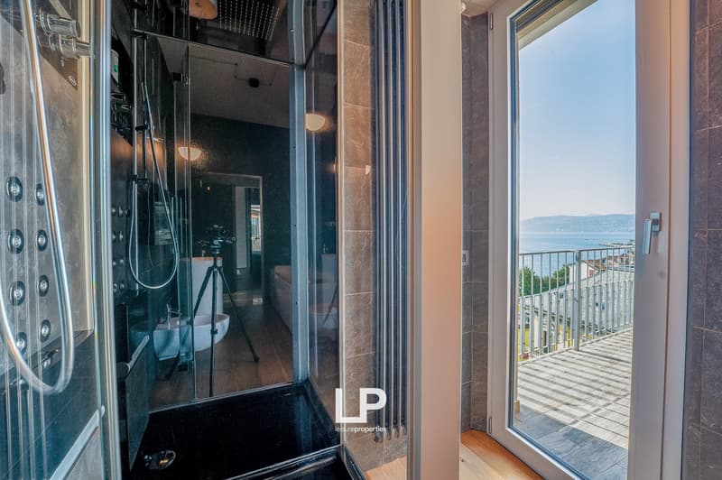 Bathroom with glass shower enclosure, tiled walls, and a view of the outdoors through a large window or sliding glass door leading to a balcony or terrace.