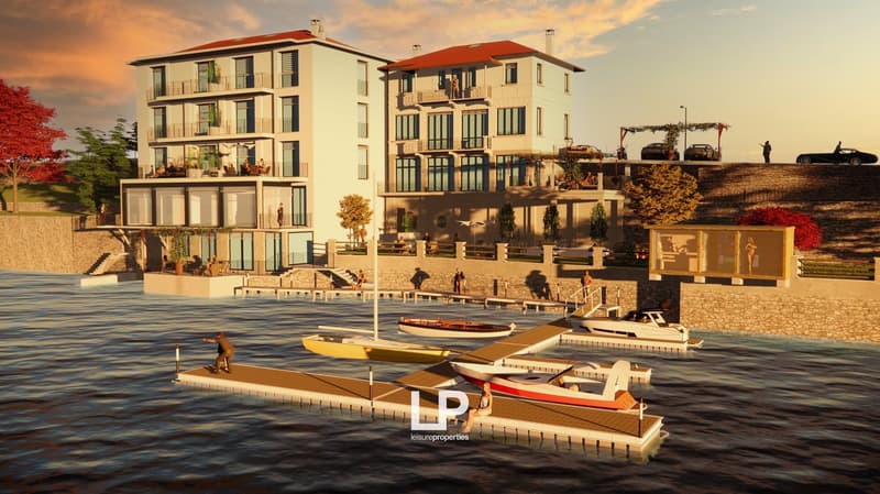 3-story waterfront buildings with multiple balconies, stone wall, boat docking area with several boats, outdoor seating area, lush greenery, distant view of mountains