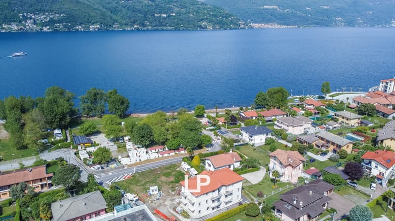 Aerial view of a lakeside town with residential buildings, greenery, and a boat on the lake. The town is surrounded by mountains and the lake appears to be a deep blue color.