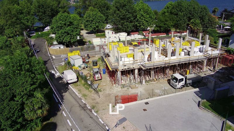 Aerial view of an active construction site with various construction materials, equipment, and vehicles visible. The site appears to be surrounded by trees and a body of water in the background.