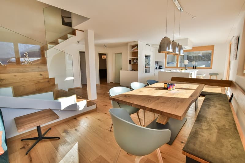 The image shows an open concept living area with a large wooden dining table and chairs, a modern kitchen with white cabinets and appliances, and a living room area with glass walls and shelving. The space has a warm, contemporary feel with hardwood floor