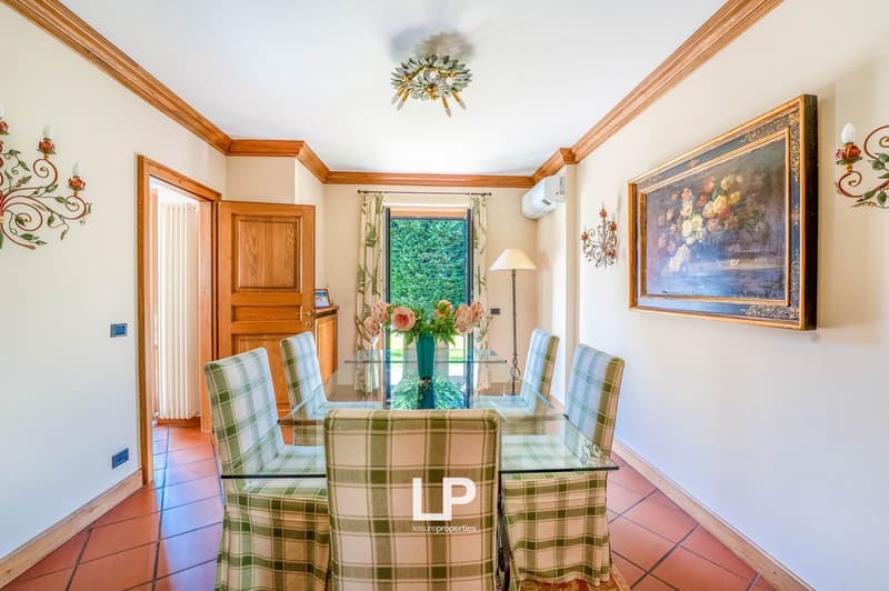 Spacious dining room with wooden trim, large windows, glass dining table, plaid upholstered chairs, and a decorative chandelier. The room also features a fireplace and a painting on the wall.