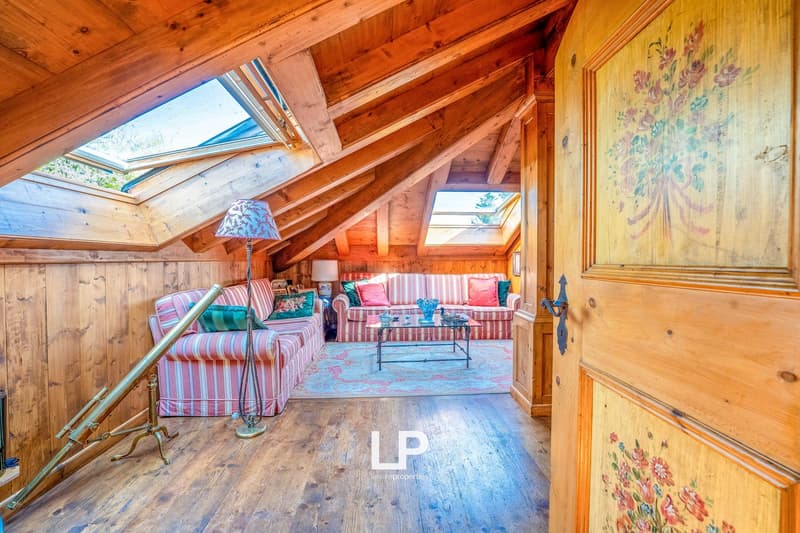 Cozy attic living room with wooden beams, skylights, floral patterned sofas, and a decorative painting on the wall
