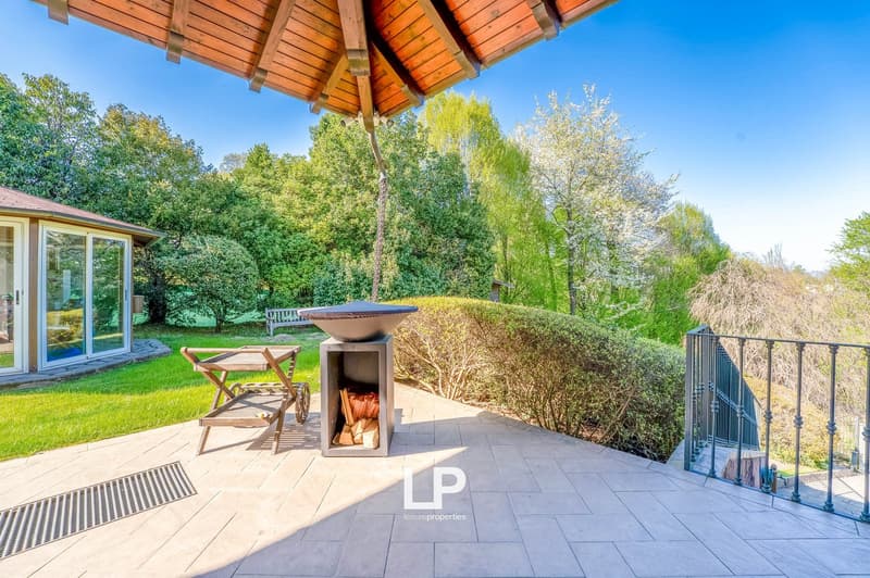 Covered patio with wooden beamed ceiling, outdoor fireplace, patio furniture, surrounded by lush greenery and trees