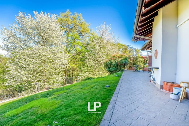 Two-story house with a patio, surrounded by lush greenery and blooming trees. The house has a tiled roof and a covered patio area with outdoor furniture. The yard features a well-manicured lawn and landscaping.