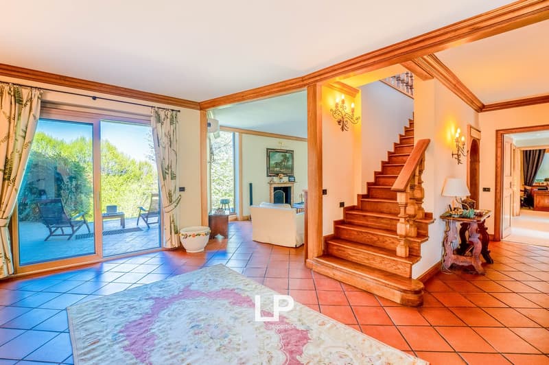 Spacious living room with large windows overlooking a balcony or terrace, hardwood floors, wooden staircase, fireplace, and decorative furnishings