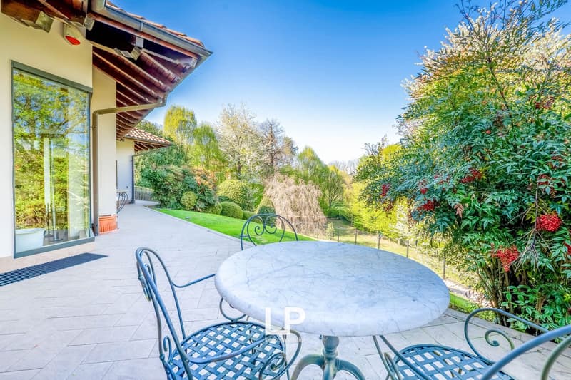 Outdoor patio with a round marble table and metal chairs, surrounded by lush greenery and trees. The house has a tiled roof and large windows overlooking the garden.
