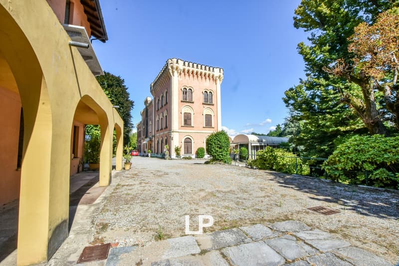 A multi-story pink and yellow castle-like building with arched windows and decorative architectural elements, surrounded by lush greenery and trees. There is a gravel driveway leading up to the building.