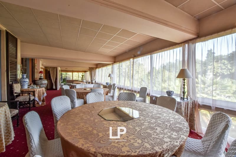 Large dining room with panoramic windows overlooking a forested area, round dining table with patterned tablecloth, upholstered chairs, curtains, and decorative lamps