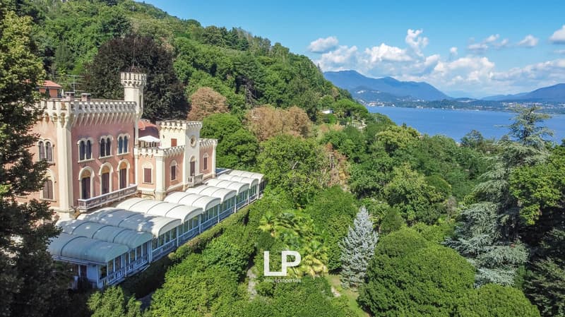 A large, ornate castle-like building with towers and arched windows, surrounded by lush green trees and vegetation overlooking a body of water. The building appears to be a historic or tourist attraction located in a scenic, mountainous area.